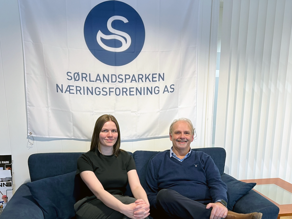 Markedskoordinator Hilde Z. Gundersen og daglig leder Erik Rostoft smiler foran logoen til Sørlandsparken Næringsforening AS.